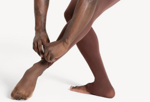 Person wearing brown tights adjusting their foot on a white background